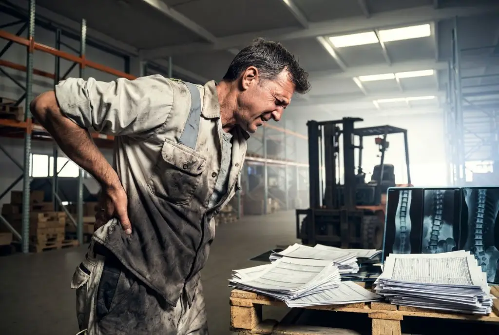 Middle-aged warehouse worker holding his lower back in pain while reviewing workers compensation medical documents and spine X-rays inside an industrial warehouse in California