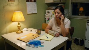 Injured California worker with a wrist brace sitting at a kitchen table at night, on the phone next to an insurance envelope, notepad, and ice pack.