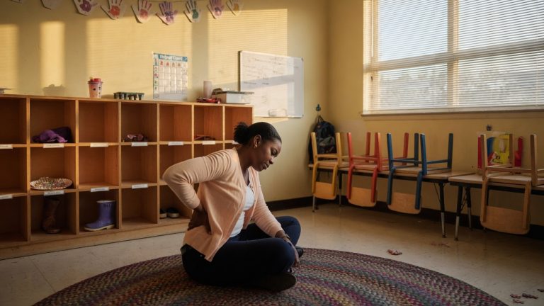 Preschool teacher sitting on a rug in an empty California classroom, holding her lower back, with small chairs and cubbies behind her.