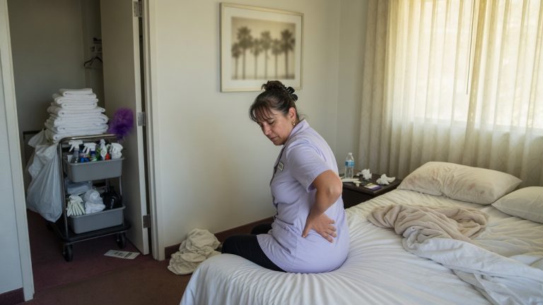 Hotel housekeeper sitting on a bed in a California hotel room, holding her lower back, with a towel cart in the doorway.