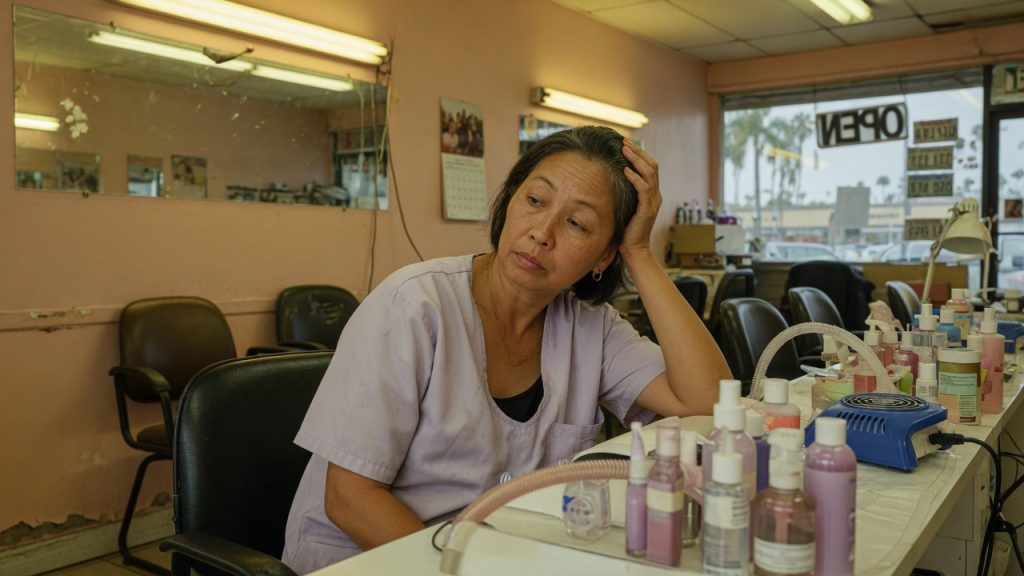 Nail technician sitting at her station in a California salon, one hand against the side of her head, with bottles and a ventilation tube on the table.