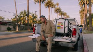 Un técnico de control de plagas sentado en el portón trasero de un camión de servicio en una calle del sur de California, con una mano sobre la rodilla, y un depósito de pulverizador en el maletero.