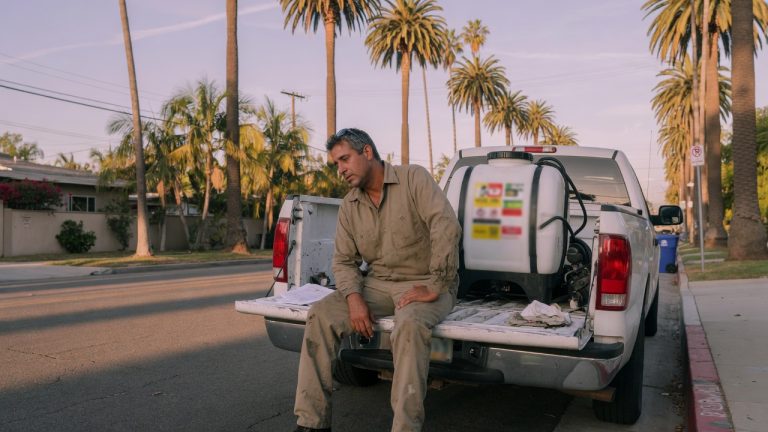 Pest control technician sitting on the tailgate of a service truck on a palm-lined Southern California street, one hand on his thigh, with a sprayer tank in the bed.