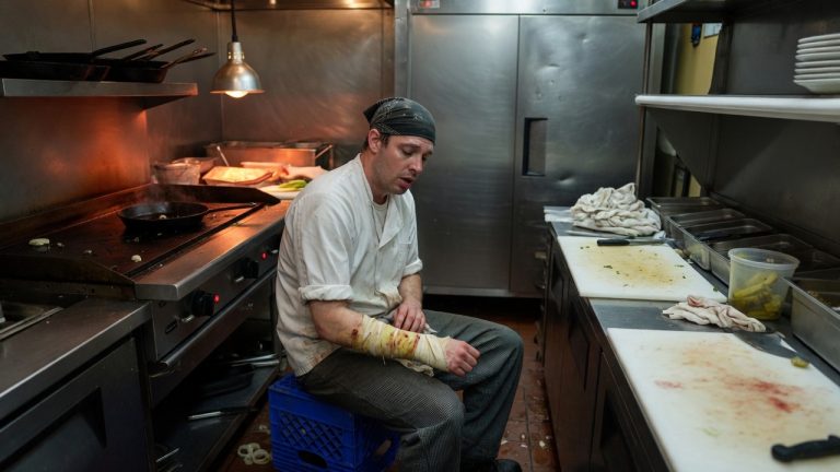 Line cook sitting on a crate in a California restaurant kitchen after close, looking at a bandaged forearm beside a flat-top grill and prep table.